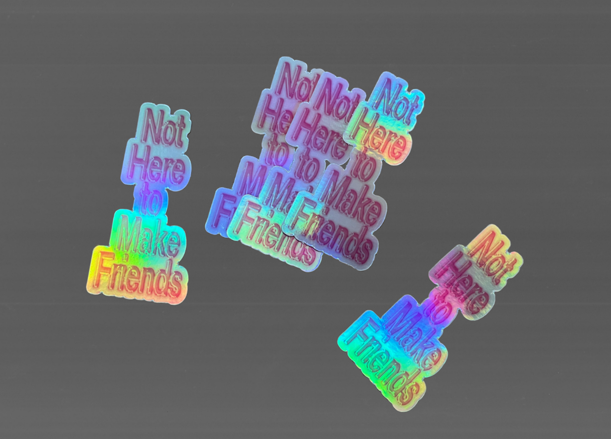 Three small holographic stickers with the text 'Not Here I Make Friends' in a rainbow iridescent finish, arranged on a plain gray background.